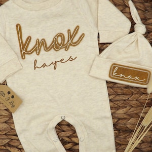 May include: A beige baby onesie with long sleeves and a brown felt applique that says "knox hayes". A matching beige hat with a brown felt applique that says "knox" is also included.