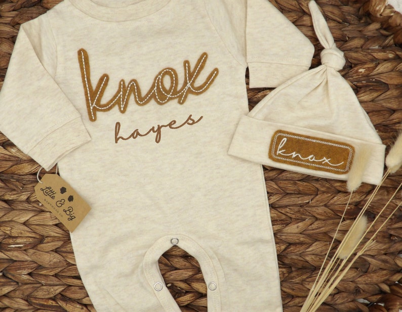 May include: A beige baby onesie with long sleeves and a brown felt applique that says "knox hayes". A matching beige hat with a brown felt applique that says "knox" is also included.