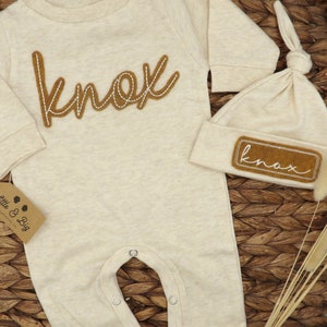 May include: A beige baby onesie with long sleeves and a brown felt appliqu&eacute; that says "knox". A matching beige beanie with a brown felt appliqu&eacute; that says "knox" is also included.