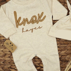 May include: A beige baby onesie with long sleeves and snaps at the crotch. The onesie has a brown felt applique that says "knox hayes". A matching beige hat is also included.