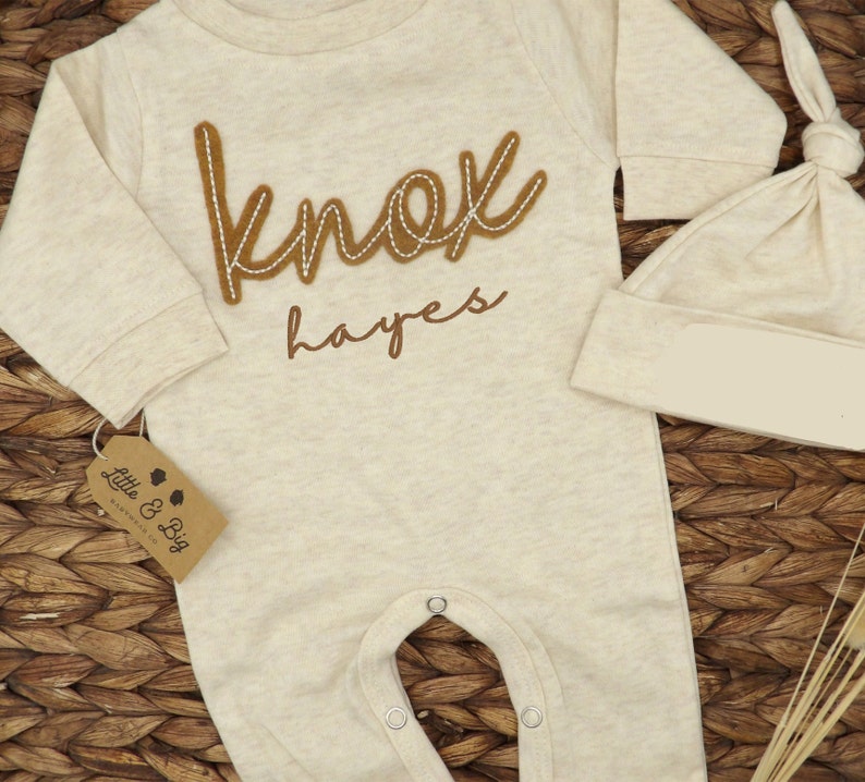 May include: A beige baby onesie with long sleeves and snaps at the crotch. The onesie has a brown felt applique that says "knox hayes". A matching beige hat is also included.