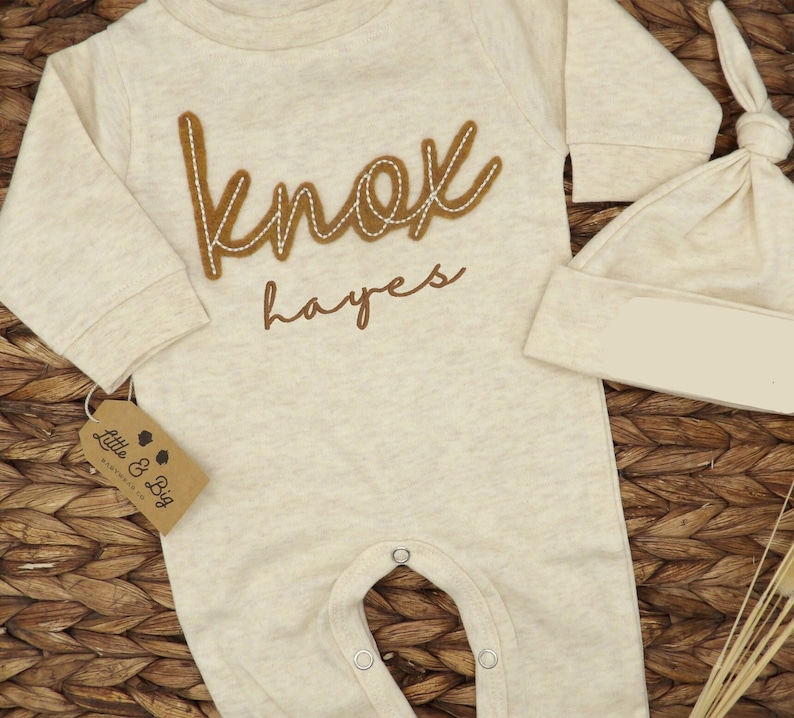 May include: A beige baby onesie with long sleeves and poppers at the crotch. The onesie has a brown felt appliqu&eacute; that says "Knox Hayes". A matching beige hat is also included.