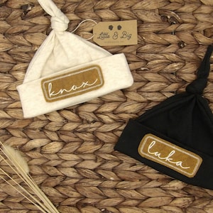 May include: Two beige and black knotted baby hats with brown fabric patches that say "knox" and "luka" in white cursive.