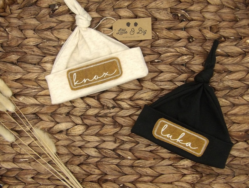 May include: Two beige and black knotted baby hats with brown fabric patches that say "knox" and "luka" in white cursive.