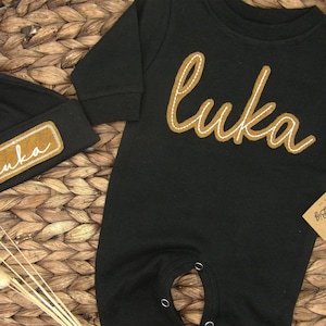 May include: Black baby onesie with long sleeves and a brown felt appliqu&eacute; of the name "Luka". A matching black beanie with a brown felt appliqu&eacute; of the name "Luka" is also included.