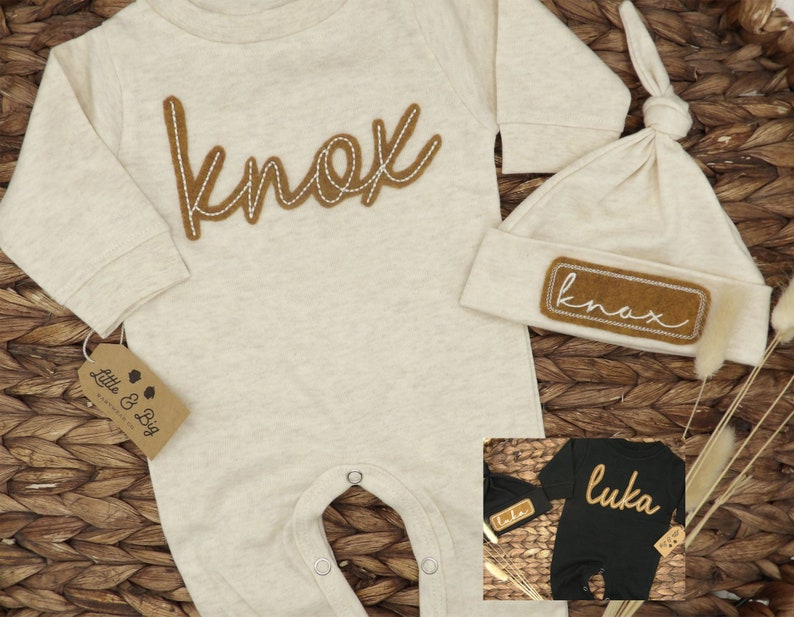 May include: A beige baby onesie with the name "knox" embroidered in brown on the front. A matching beige hat with the name "knox" embroidered in brown on a patch is included. The onesie and hat are on a woven brown background.