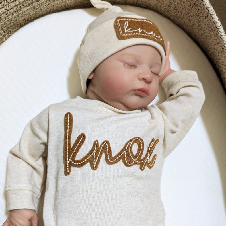 May include: A newborn baby wearing a cream-colored long-sleeve onesie with a brown felt "Knox" applique. The baby is also wearing a cream-colored knit hat with a brown felt "Knox" applique.