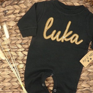 May include: Black long-sleeve baby onesie with a brown embroidered name "Luka" on the front. The onesie has snaps at the crotch and a tag that says "Little & Big".