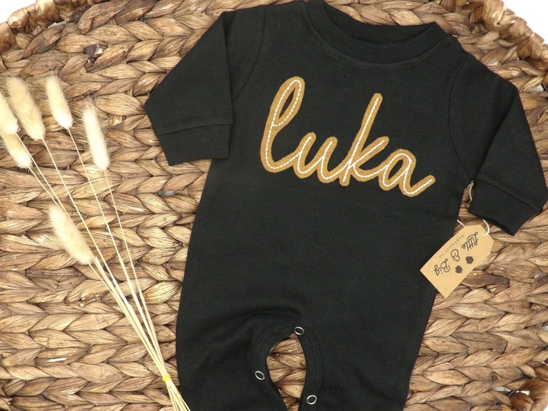 May include: Black long-sleeve baby onesie with a brown embroidered name "Luka" on the front. The onesie has snaps at the crotch and a tag that says "Little & Big".