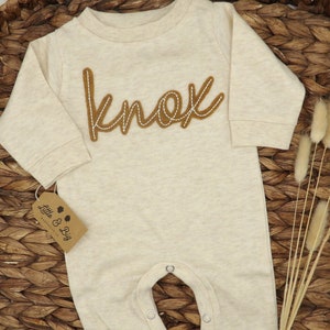 May include: A beige long-sleeve baby onesie with the name "Knox" embroidered in brown on the front. The onesie has snaps at the crotch and a tag that says "Little & Big" on the bottom left side.