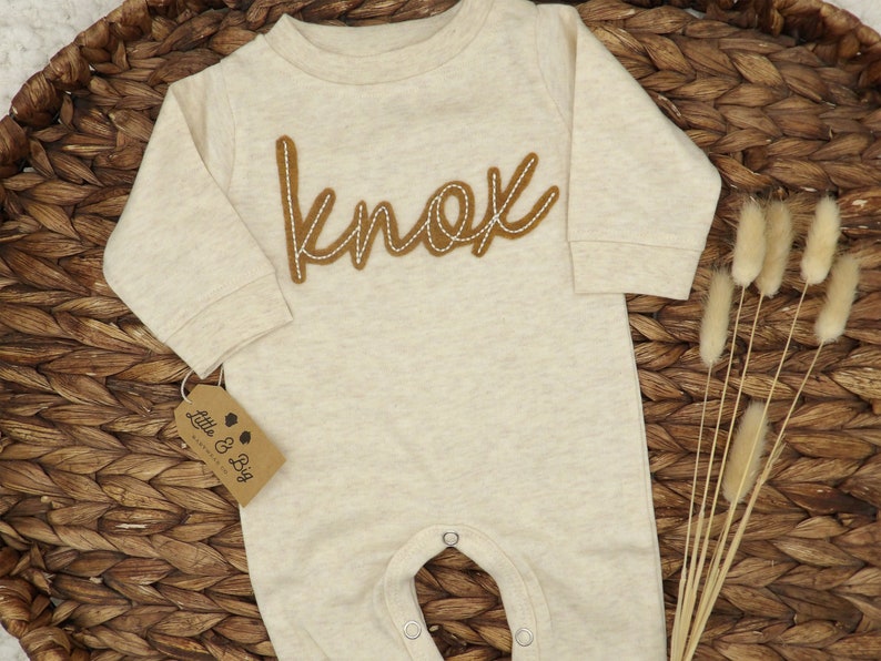 May include: A beige long-sleeve baby onesie with the name "Knox" embroidered in brown on the front. The onesie has snaps at the crotch and a tag that says "Little & Big" on the bottom left side.