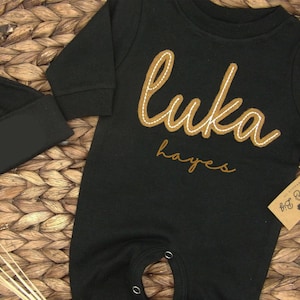 May include: Black baby onesie with long sleeves and brown embroidered text that says "Luka Hayes".
