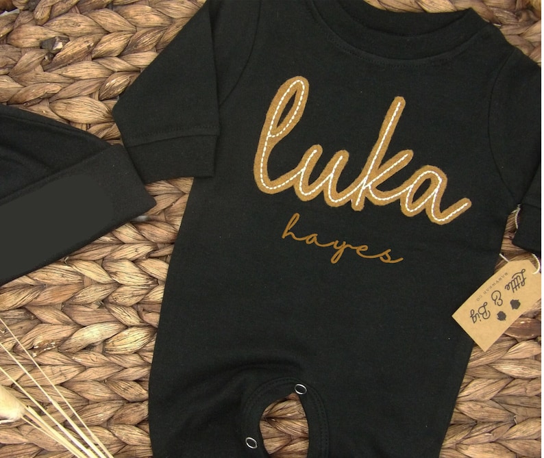 May include: Black baby onesie with long sleeves and brown embroidered text that says "Luka Hayes".