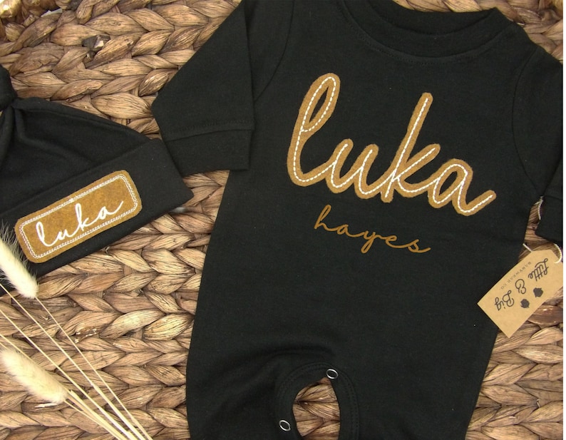 May include: Black baby onesie with long sleeves and a brown felt patch with the name "Luka Hayes" in white stitching. A matching black beanie with a brown felt patch with the name "Luka" in white stitching.