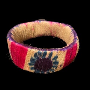 May include: A woven bangle bracelet with a pink, white, and blue pattern. The center of the bracelet features a purple and blue floral design.
