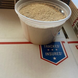 May include: A clear plastic container filled with a light brown powder. The container is sitting on a white surface with a blue and red "Tracked & Insured" sticker.