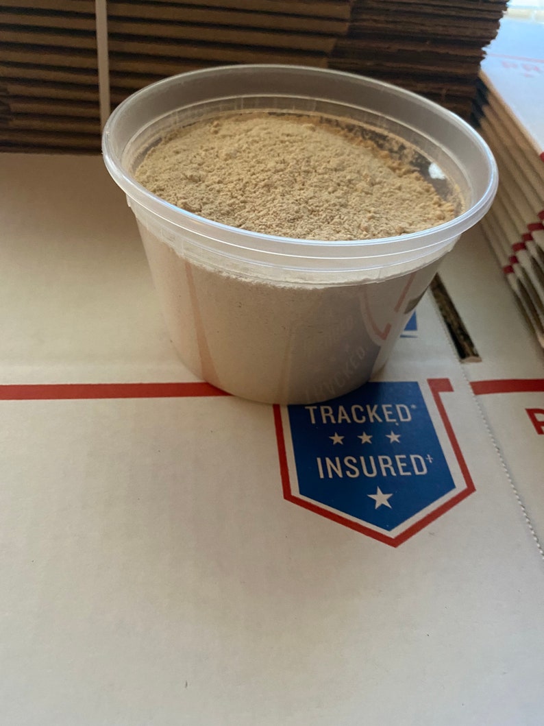 May include: A clear plastic container filled with a light brown powder. The container is sitting on a white surface with a blue and red "Tracked & Insured" sticker.