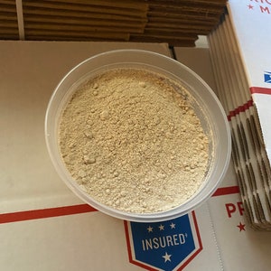 May include: A clear plastic container filled with a light beige powder. The container is sitting on a white surface with a red line and a blue and white sticker that says "INSURED".