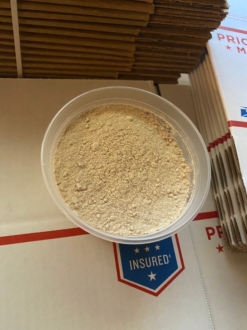 May include: A clear plastic container filled with a light beige powder. The container is sitting on a white surface with a red line and a blue and white sticker that says "INSURED".