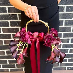 May include: A gold-coloured metal handle supports a bouquet of deep burgundy calla lilies and other flowers. A matching burgundy ribbon is tied in a bow and hangs down. The flowers are arranged in a circular shape.