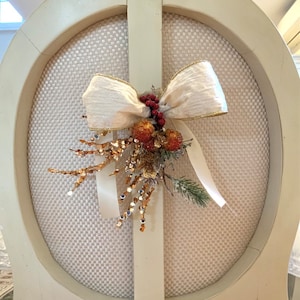 May include: A cream-coloured chair back adorned with a decorative bow and floral arrangement. The bow is made of a sheer, gold-trimmed fabric. The arrangement includes red berries, gold-toned sprigs, and a small pine branch, creating a festive look.