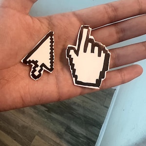 May include: Two white paper stickers with black outlines of a computer mouse cursor and a hand pointing. The stickers are on a person's hand.
