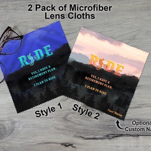 May include: Two microfiber lens cloths with a mountain biking theme. One cloth is blue with a turquoise design and the other is orange with a sunset background. Both cloths feature the text "RIDE" and the phrase "Yes, I have a retirement plan. I plan to ride."