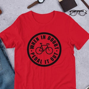 Könnte beinhalten: Rotes T-Shirt mit einem schwarzen Kreis-Motiv, auf dem "When in doubt, pedal it out." steht. Das Motiv enthält auch eine Silhouette eines Fahrrads.
