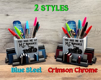 Könnte beinhalten: Zwei 3D-gedruckte Stifthalter in Form eines Bücherstapels. Das vordere Buch trägt den Text "You Had Me At M.C. Romance" und eine Motorradgrafik. Ein Halter ist aus blauem Stahl, der andere aus rotbraunem Chrom.
