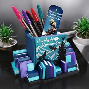 Peut inclure: Un porte-stylos en forme de livre bleu et turquoise avec un motif de loup et de dragon. Le livre est intitulé "In My Shifter Romance Era". Le porte-stylos est entouré de piles de livres colorés.