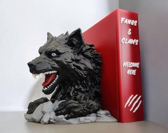 May include: A black wolf bookend with a snarling expression, white fangs, and a red mouth. The bookend is paired with a red book with the text "FANGS & CLAWS WELCOME HERE" in white. The bookend is on a grey base.