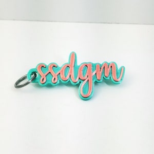 Peut inclure: Un porte-clés turquoise et corail avec les lettres "ssdgm" dans une police cursive. Le porte-clés est muni d'un anneau en métal argenté. Les lettres sont de couleur corail et le fond est turquoise.