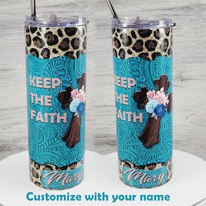 May include: Two stainless steel tumblers with a turquoise background and a brown cross with pink and blue flowers. The tumblers have a leopard print design on the bottom and the words "Keep the Faith" on the front. The name "Mary" is written on the bottom of each tumbler.