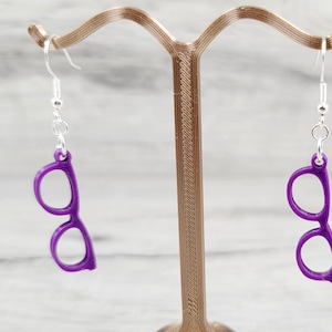 May include: A pair of purple eyeglasses-shaped earrings hanging from silver hooks. The earrings are a solid, vibrant purple color and are displayed on a decorative stand. The earrings are a fun and unique accessory.