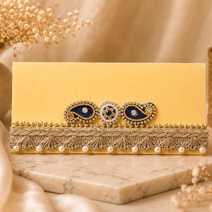 May include: A rectangular pale yellow envelope featuring a horizontal decorative band of metallic gold lace trim with small white pearls. The center is adorned with two dark blue paisley motifs and a central circular floral embellishment.