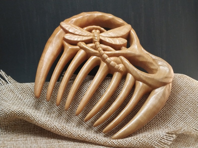 Puede incluir: Un peine de madera con un dise&ntilde;o de lib&eacute;lula tallado. El peine tiene siete dientes y est&aacute; hecho de madera marr&oacute;n claro.