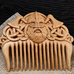 May include: A light brown wooden comb featuring a detailed carving of a Viking warrior wearing a horned helmet, flanked by two crossed axes and intricate Celtic knotwork patterns. The comb has eleven sturdy teeth and a smooth, polished finish.