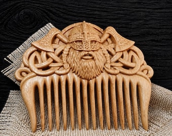 May include: A light brown wooden comb featuring a detailed carving of a Viking warrior wearing a horned helmet, flanked by two crossed axes and intricate Celtic knotwork patterns. The comb has eleven sturdy teeth and a smooth, polished finish.
