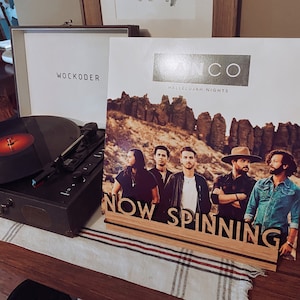 May include: A record player with a black vinyl record on the turntable. The record player is on a wooden table with a white and black striped tablecloth. A record album cover is leaning against the record player. The album cover is white with the text "Hallelujah Nights" and a band photo. The band is standing in front of a rocky landscape. The album cover also has the text "Now Spinning" on a wooden sign.