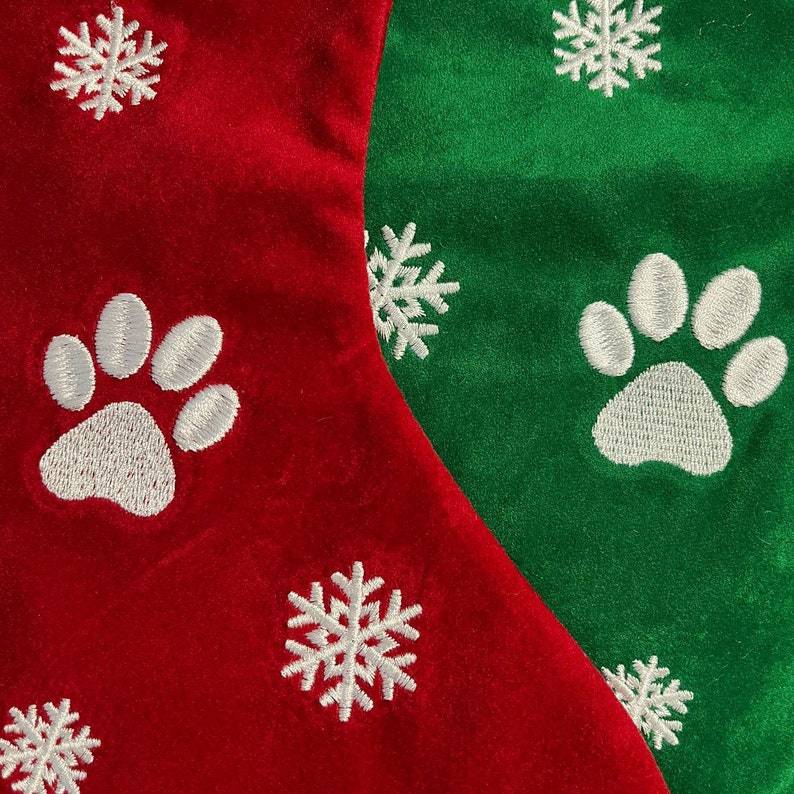 May include: Close-up of two Christmas stockings, one red and one green. Each stocking features white embroidered paw prints and snowflakes. The stockings are made of a soft, velvety material, perfect for pet lovers.