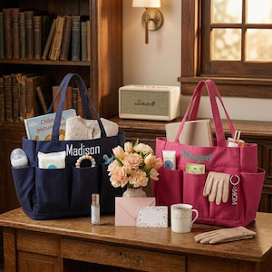 May include: Two tote bags, one navy blue and one pink, sit on a wooden table. The navy bag is labeled "Madison" and contains baby items. The pink bag, labeled "Margaret", holds personal care items. A vase of flowers and other accessories are also on the table.
