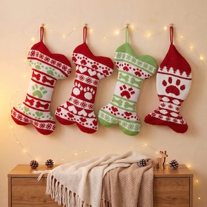 May include: Four knitted stockings hanging in a row. Three are shaped like dog bones and one is shaped like a fish. They feature red, white, and green patterns including paw prints, hearts, and bones. Each stocking has a loop for hanging.