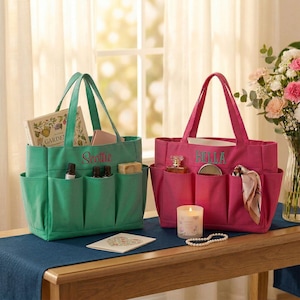 May include: Two tote bags, one teal and one fuchsia, sit on a wooden table. The teal bag has "Scottie" embroidered on it and contains a book, essential oils, and soap. The fuchsia bag has "Bella" embroidered and holds perfume and a scarf.