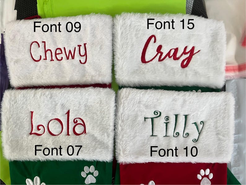 May include: Four Christmas stockings with white faux fur cuffs and red and green bodies. Each stocking has a name embroidered in a different font: "Chewy", "Cray", "Lola", and "Tilly". Paw prints decorate the bottom of the stockings.