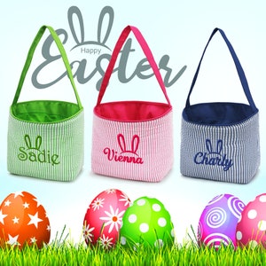 May include: Three personalized Easter baskets with striped fabric and bunny ears embroidery. The baskets are green, pink, and blue. The names Sadie, Vienna, and Charly are embroidered on the baskets. The baskets are sitting on a bed of green grass with colorful Easter eggs.