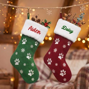 May include: Two Christmas stockings with white faux fur cuffs. One is green with the name Aiden in red embroidery, the other is red with the name Daisy in green embroidery. Both feature white paw prints and snowflake patterns throughout.
