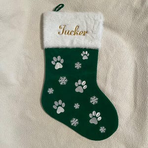May include: A green Christmas stocking with white paw prints and snowflakes embroidered on the front. The top of the stocking is white faux fur, with the name "Tucker" embroidered in gold thread.