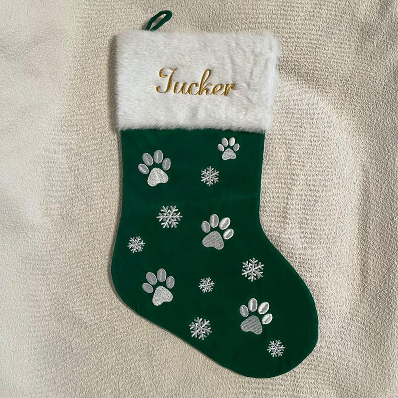 May include: A green Christmas stocking with white paw prints and snowflakes embroidered on the front. The top of the stocking is white faux fur, with the name "Tucker" embroidered in gold thread.