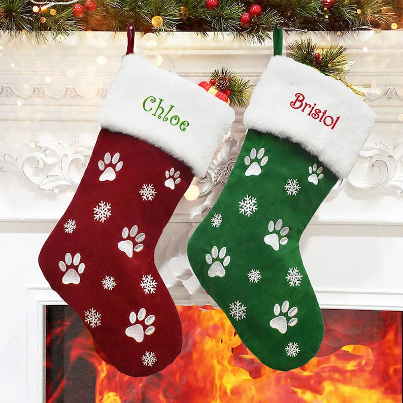 May include: Two Christmas stockings, one burgundy and one green, hang in front of a fireplace. The burgundy stocking has the name "Chloe" embroidered in green, and the green stocking has "Bristol" embroidered in red. Both stockings feature paw prints and snowflakes.