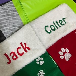 May include: Christmas stockings with white faux fur cuffs. One green stocking has the name "Jack" embroidered in red, and paw prints. Another green stocking has the name "Colter" embroidered in green. A third red stocking has paw prints.